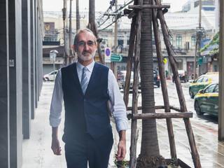 business man senior in formal suit stand confidently with ooking ahead on city street. Modern building, palm trees, and urban element in background create professional and metropolitan atmosphere.