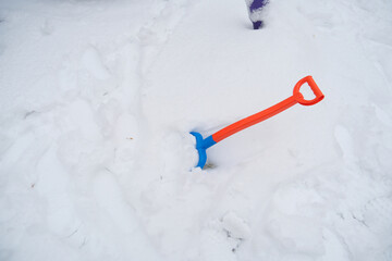 An orange and blue shovel is partially buried in deep white snow, indicating recent snowfall. The vibrant colors contrast with the stark white snow, emphasizing winter conditions.