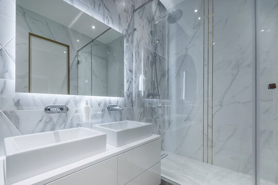 A bathroom with marble walls and bright lights. Two sinks are on a cabinet. The glass shower is enclosed and has a sleek look. The room has a clean feel