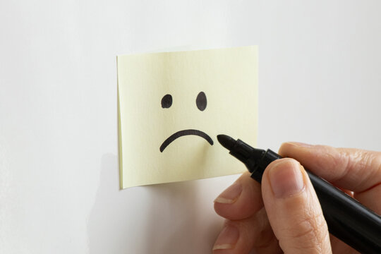 A yellow sticky note with a drawn sad face and a woman's hand holding a black marker on a white background, depicting bad mood, sadness