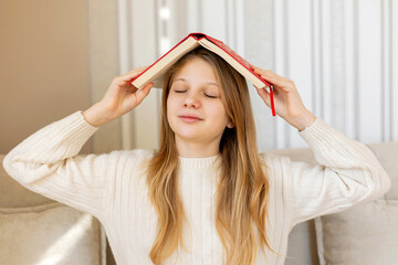 Smiling teen girl relaxing with book on head. A teenage girl playfully balances a book on her head while smiling with closed eyes, enjoying a peaceful and carefree moment indoors.
