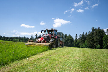 Fototapeta premium Grünfutterernte im Allgäu, Traktor mit vorgebautem Mähwerk und Ladewagen bei der Grasernte.