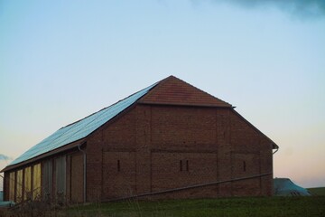 Alte rotbraune Backsteinscheune mit Solardach auf gr&uuml;nem Ackerfeld vor hellblauem Himmel auf dem Land  bei Sonnenuntergang am Abend im Winter