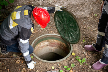 Two workers near an open manhole. A worker in a red hard hat leans over to open and inspect the round manhole, peering into the underground shaft of a septic tank