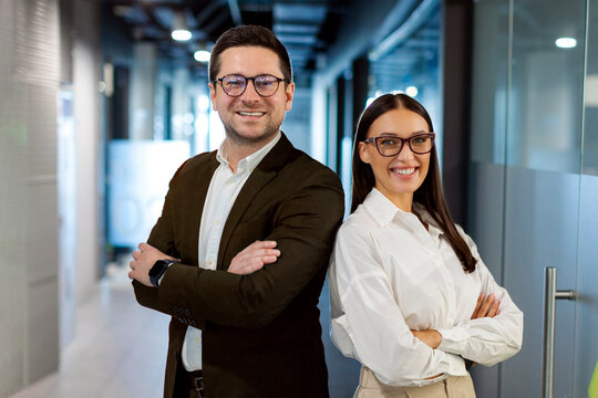 Positive European business partners posing for portrait at office hallway interior standing back to back with arms crossed, looking at camera and smiling