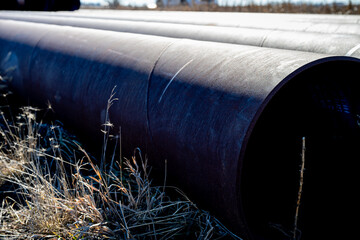 Large black pipes lie in a field near industrial buildings during a clear day