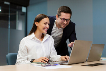 Fototapeta premium Positive European businesspeople discussing goals and plans during meeting together in modern office, sitting at desk and using laptop