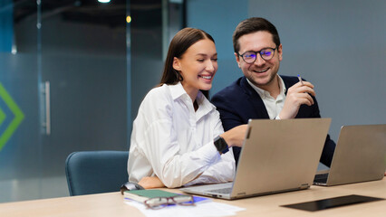 Two positive colleagues engaging in discussion, using laptop for project collaboration in modern office environment, sitting at desk in board room