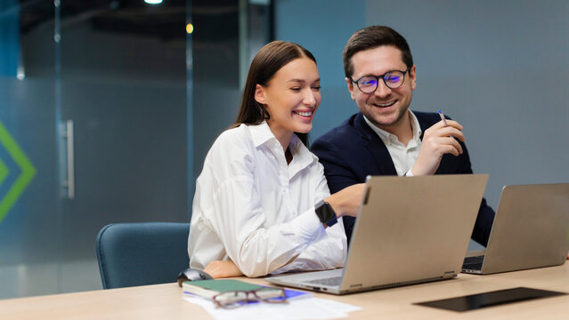 Two positive colleagues engaging in discussion, using laptop for project collaboration in modern office environment, sitting at desk in board room - Powered by Adobe