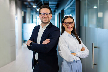 Positive European business partners posing for portrait at office hallway interior standing back to back with arms crossed, looking at camera and smiling