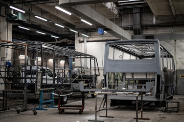Industrial vehicle manufacturing workshop with metal bus frames and body structures under assembly. Large factory interior shows fabrication, welding, and custom vehicle production in progress.