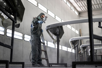 Worker spray painting vehicle body part in professional paint booth. Industrial automotive painting process with protective equipment, precision tools, and controlled environment.