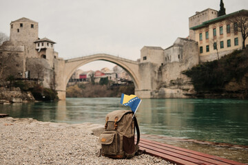 Backpack with Bosnia and Herzegovina flag placed on riverbank near Stari Most bridge in Mostar. Concept of travel, backpacking, freedom and exploration of Balkan heritage.