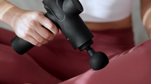 Female using a vibrating massage gun on her thigh while seated on a soft surface, showcasing the massage tool's application and effectiveness in muscle relaxation and recovery