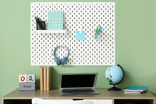 Comfortable workplace with modern laptop, stationery, globe and pegboard hanging on color wall
