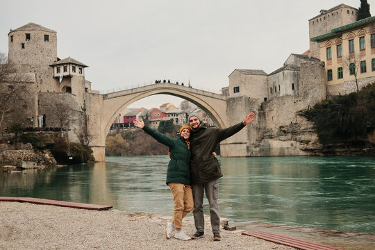 Smiling couple with open arms standing by the river near the historic bridge in Mostar. Travel freedom and shared happiness concept inspired by exploration and cultural discovery.