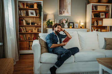 Thoughtful man relaxing reading book on sofa at home library