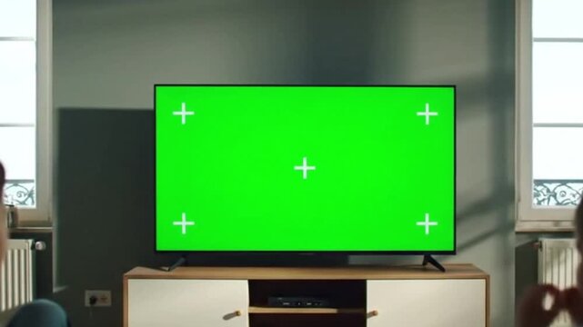 A modern flat-screen television with a vibrant green screen and four white corner markers, positioned on a minimalist wooden media console in a well-lit living room with large windows in the backgro