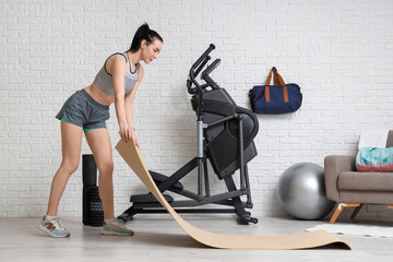 Sporty young woman placing yoga mat near modern exercising bike in stylish gym