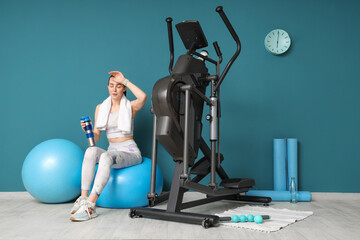 Sporty young woman with water bottle resting after training on modern exercising bike in stylish gym