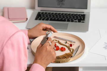 Young African-American female jewelry designer examining gemstones and necklace with magnifier in workshop, closeup