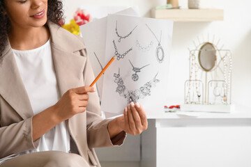 Young African-American female jewelry designer pointing at drawings of adornments in workshop,...