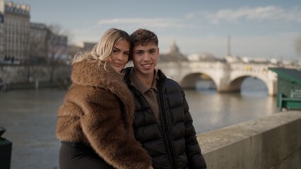 A young couple poses for a portrait by the Seine River in Paris France on a sunny day The cityscape and bridges provide a romantic backdrop for tourism and travel related content.