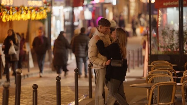 A romantic couple embraces on a Parisian street at night surrounded by tables and festive Christmas lights. This image captures the essence of love and connection in the heart of France suitable for t