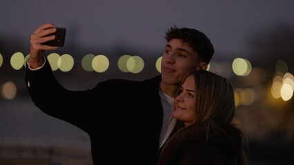 A young couple takes a selfie with a smartphone in Paris France at night with bokeh lights in the background. This image represents love connection travel and tourism.
