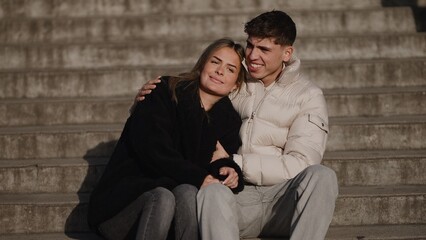 A young couple in love sits on concrete steps in Paris France wearing winter coats and jeans They are enjoying a romantic moment together outdoors in the city.