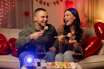 Happy man and woman enjoy eating sushi, using chopsticks and laughing, having romantic date and dinner in room decorated with hearts and warm lighting