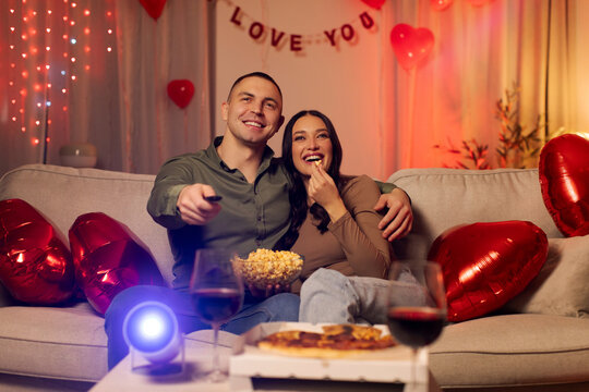 Loving couple having movie night at home, during romantic date, . eating popcorn and pizza while watching film on projector, room filled with red heart balloons for Valentine's Day