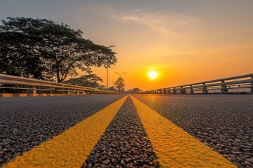 Low Angle View of Asphalt Road with Yellow Lines Leading to Golden Sunset Under Cloudy Sky and Silhouetted Trees in Background