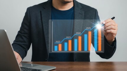 A businessman in a dark suit points to a glowing holographic bar graph depicting upward trends and growth predictions from 2022 to 2026 with a laptop visible on the desk chart statistics