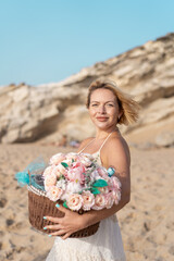 Smiling young woman in dress holding basket with pink flowers on sandy beach enjoying summer vacation near rocky cliffs. Beautiful lady with short blond hair looking at camera