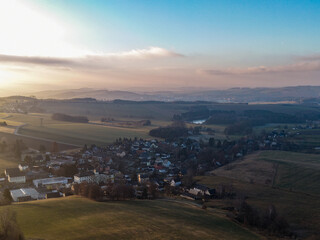 calm village rests in a wide valley surrounded by rolling hills, bathed in soft sunlight with misty layers fading into the distant landscape