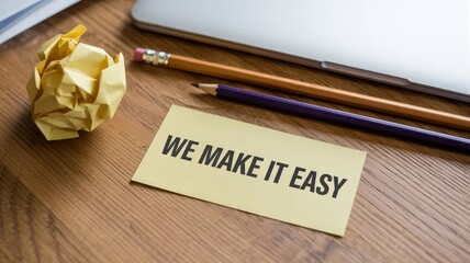 Yellow sticky note displaying the motivational phrase we make it easy rests near crumpled paper and pencils on a wooden desk.