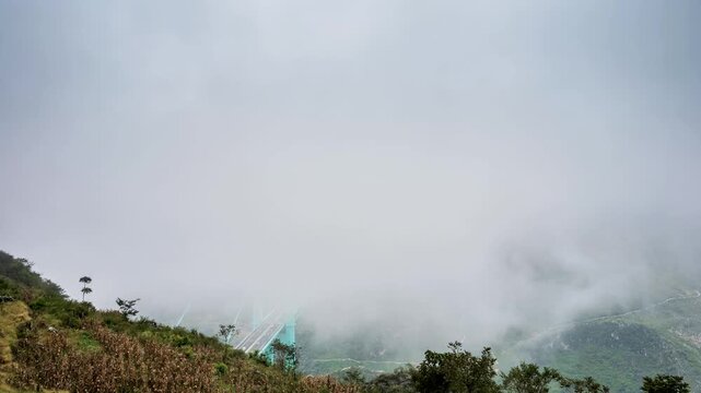 time-lapse of the Huajiang Canyon Bridge in Guizhou, China, emerging from thick mist over a deep canyon. Dramatic clouds swirl around the modern suspension bridge.
