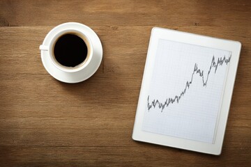 Coffee and tablet display financial chart on wooden table in morning hours near window