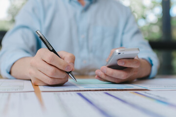 Close-up of businessman holding smartphone and writing on paper document, multitasking during data review or financial analysis in office workspace Modern business communication concept.