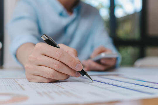 Close-up of businessman holding smartphone and writing on paper document, multitasking during data review or financial analysis in office workspace Modern business communication concept. - Powered by Adobe