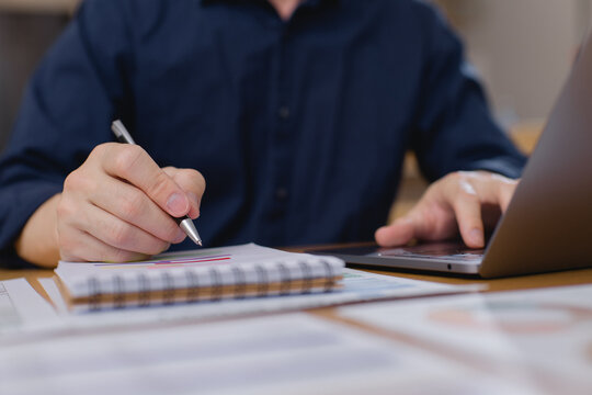 businessman writing in notebook while using laptop, taking notes from financial data and business reports in office environment. Productivity and planning concept.