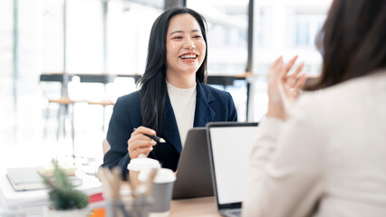 Two professional Asian women collaborating and reviewing documents on a desk in a modern office workspace.