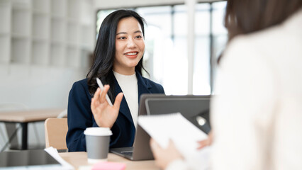 Confident Asian businesswoman smiling and discussing work with laptop and notebook in a modern coworking space.