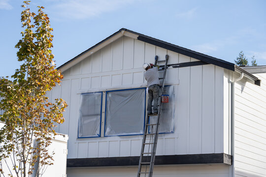 Professional house painter on an extension ladder performing detail trim work with paintbrush on new home in residential development, housing growth
