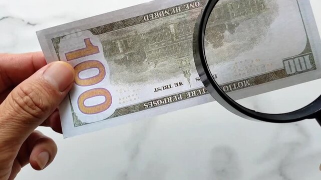 Counterfeit money. A man examines a hundred-dollar bill with a magnifying glass. Looking for watermarks on the bill. The concept of counterfeit money exchange.