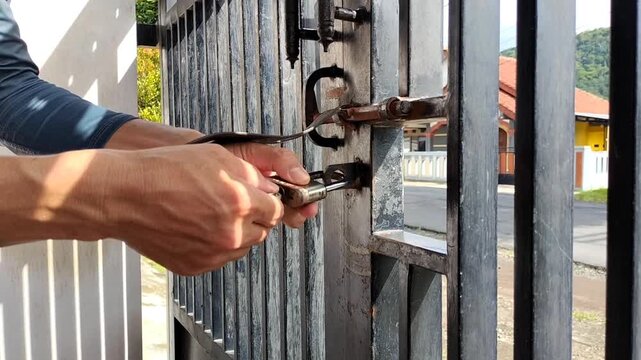 Kuningan City, Indonesia, December 25, 2025: A man's hand locks an iron padlock with a key. The black-painted gate looks a little worn. A security concept.