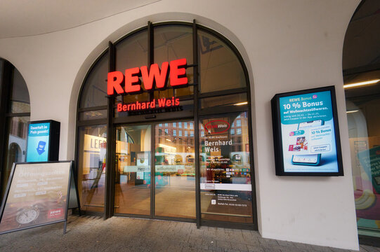 Cologne, Germany December 22 2025: Illuminated entrance of REWE Bernhard Weis supermarket in Cologne Braunsfeld, Germany. Modern retail architecture with arched windows and digital signage