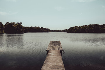 A wooden dock stretches over calm water beneath a clear sky surrounded by distant green trees the peaceful scene feels open tranquil and inviting in natural daylight