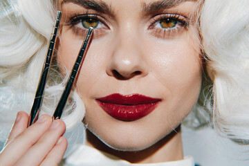 Elegant woman with striking makeup holding tweezers, showcasing beauty artistry and vibrant emotions against a minimalist background.